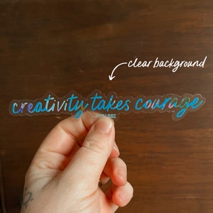 K&ouml;nnte beinhalten: Ein transparenter Aufkleber mit dem Text "creativity takes courage" in einem blauen und violetten Farbverlauf. Der Aufkleber befindet sich auf einem Holzgrund.