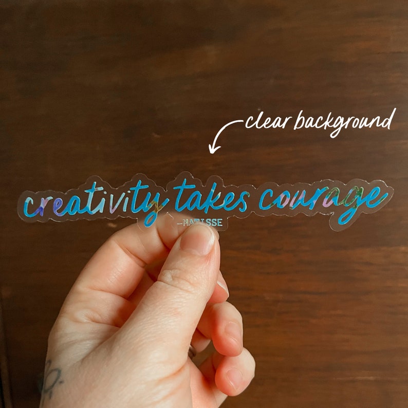 K&ouml;nnte beinhalten: Ein transparenter Aufkleber mit dem Text "creativity takes courage" in einem blauen und violetten Farbverlauf. Der Aufkleber befindet sich auf einem Holzgrund.