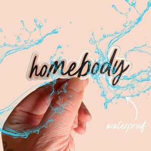 Pu&ograve; includere: Un adesivo rosa e nero con il testo "homebody" in un font di scrittura a mano. L'adesivo &egrave; impermeabile ed &egrave; tenuto da una mano.