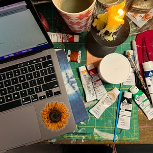 Peut inclure: Un espace de travail avec un ordinateur portable, du mat&eacute;riel d'art et une bougie allum&eacute;e. L'ordinateur portable a un autocollant de tournesol. Des tubes de peinture Gouache de diff&eacute;rentes couleurs, un pinceau et une tasse de liquide sont sur un tapis de coupe vert.