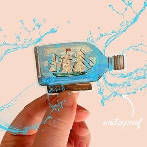 May include: A clear glass bottle with a cork stopper containing a miniature ship. The bottle is surrounded by blue water droplets, indicating it is waterproof.