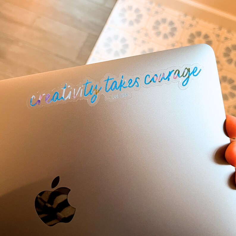 K&ouml;nnte beinhalten: Ein silberner Laptop mit einem durchsichtigen Aufkleber, auf dem "creativity takes courage" steht. Der Aufkleber ist in Blau, Rosa und Gr&uuml;n gehalten.