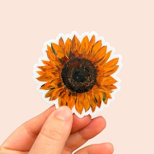 May include: A vibrant sticker featuring a detailed painting of a sunflower. The sunflower has bright yellow-orange petals surrounding a dark brown center. The sticker has a white border and is held against a light pink background.