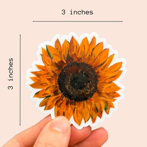 Peut inclure: Un autocollant de tournesol de 7,6 cm avec un motif aquarelle. L'autocollant pr&eacute;sente une repr&eacute;sentation d&eacute;taill&eacute;e d'un tournesol avec des p&eacute;tales jaunes et un centre brun fonc&eacute;. L'autocollant est tenu par une main sur un fond rose clair.