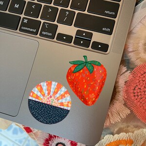 May include: Two colorful fabric patches on a silver laptop. One patch is a red strawberry with green leaves and white and yellow floral details. The other patch is a blue and orange sun with a black and white wave design.