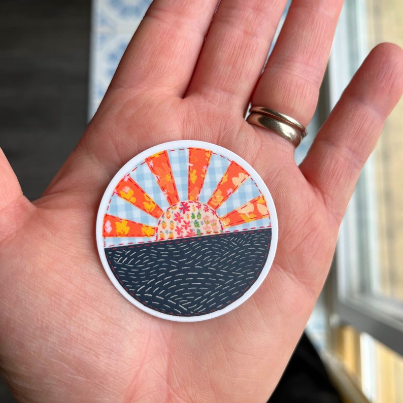 May include: A round, white sticker with a colorful, patchwork-style design of a sunset over a dark blue sea. The sunset is made of orange, red, and yellow fabric with a white and blue gingham pattern. The sea is made of dark blue fabric with white stitching.