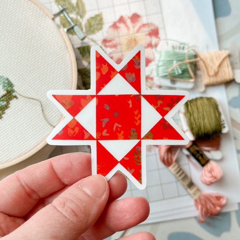 May include: A red and white star-shaped sticker with a quilt block design. The sticker features a floral pattern within the red sections. Embroidery floss and a hoop are visible in the background.