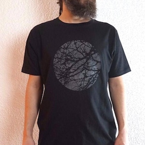 Puede incluir: Camiseta negra con un diseño circular gris de ramas de árboles. La camiseta de manga corta se combina con pantalones negros. La persona de la imagen está de pie frente a una pared blanca texturizada.