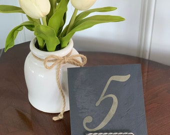 May include: A black slate table number with the number 5 written in gold paint. The number is on a silver stand and is next to a white vase with white tulips.