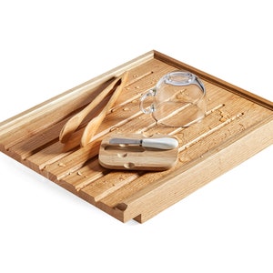 May include: A wooden dish drying rack with a rectangular shape and grooved surface. It holds two wooden spoons, a small bamboo cutting board with a butter knife, and a clear glass teacup. Water droplets are visible on the rack.