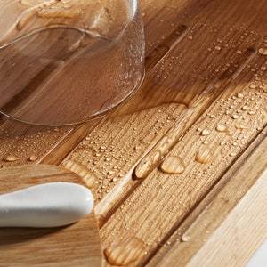 May include: Close-up of a wooden bath tray with a glass and a white ceramic object. The tray has a light brown finish with water droplets scattered across the surface. The glass is partially visible, and the ceramic object is smooth and curved.