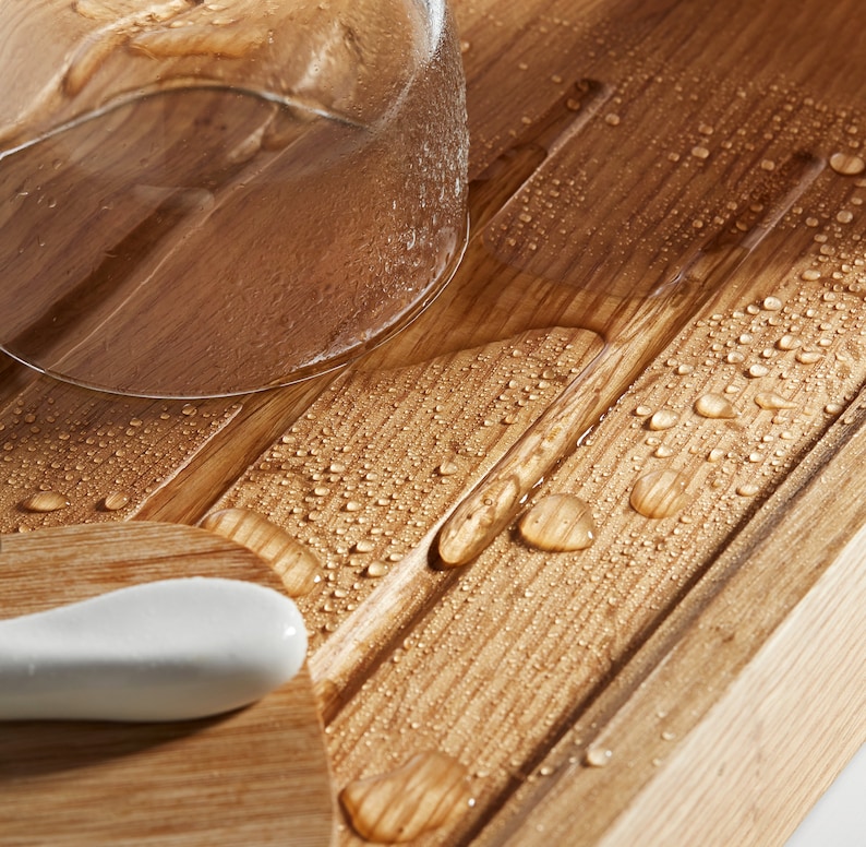 May include: Close-up of a wooden bath tray with a glass and a white ceramic object. The tray has a light brown finish with water droplets scattered across the surface. The glass is partially visible, and the ceramic object is smooth and curved.