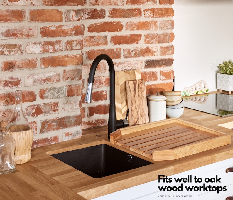 May include: A kitchen sink with a black faucet and a wooden drying rack. The sink is set into a light oak countertop, with a brick wall in the background. The text "Fits well to oak wood worktops" is visible.