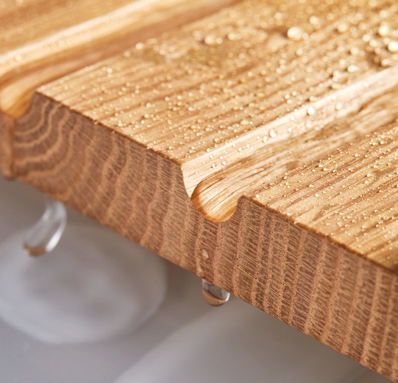 May include: Close-up of a wooden bath mat with water droplets. The light brown wood has a grooved design and rounded edges. Water beads on the surface and drips off the side, creating a natural and spa-like aesthetic.
