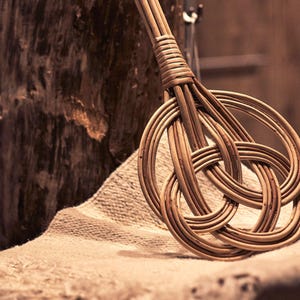 May include: A woven wicker whisk with a knotted handle. The whisk is made of natural brown wicker and is lying on a beige fabric surface.