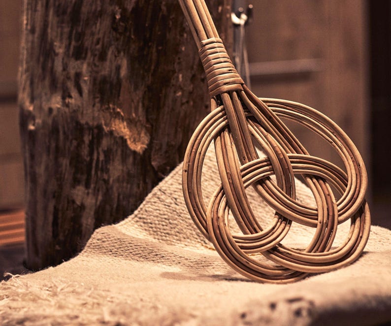 May include: A woven wicker whisk with a knotted handle. The whisk is made of natural brown wicker and is lying on a beige fabric surface.