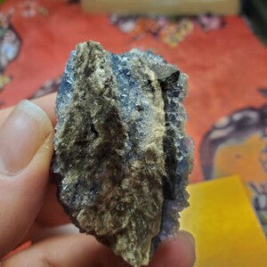 May include: A rough, natural mineral specimen with a mix of blue and tan colors. The blue crystals are clustered together, while the tan areas have a rough, earthy texture. The mineral is held in a hand.