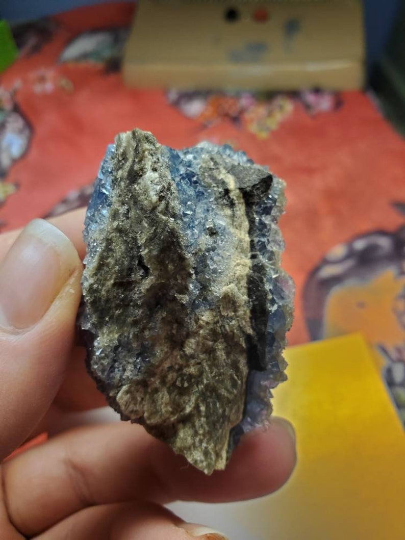 May include: A rough, natural mineral specimen with a mix of blue and tan colors. The blue crystals are clustered together, while the tan areas have a rough, earthy texture. The mineral is held in a hand.
