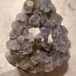 May include: A cluster of blue-purple fluorite crystals. The crystals are cubic in shape and are set in a matrix of dark brown material. The fluorite has a translucent appearance, with some areas appearing more opaque. The overall shape is irregular.