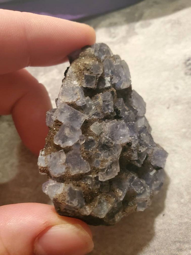 May include: A cluster of light purple fluorite crystals with a cubic structure. The crystals are set in a matrix of brown and gray rock. The image is a close-up, highlighting the mineral's texture and color.