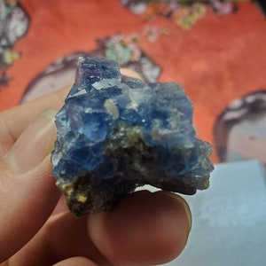 May include: A blue fluorite crystal cluster with a rough, natural texture. The crystal displays shades of blue and is partially embedded in a matrix of darker rock. The crystal is held in a hand.