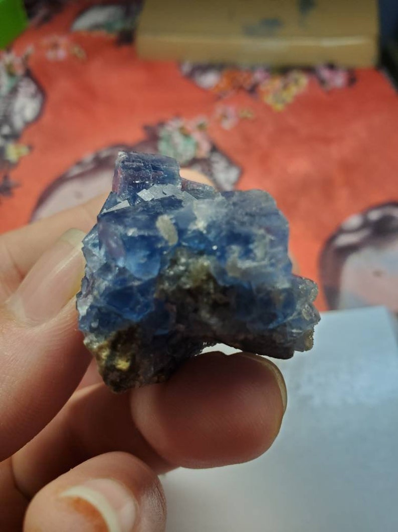 May include: A blue fluorite crystal cluster with a rough, natural texture. The crystal displays shades of blue and is partially embedded in a matrix of darker rock. The crystal is held in a hand.