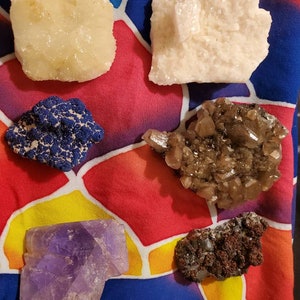 May include: Assortment of raw crystals and minerals, including a large, light purple amethyst, a dark blue cluster, and several other specimens in shades of white, brown, and yellow. The crystals are displayed on a colorful, patterned fabric.