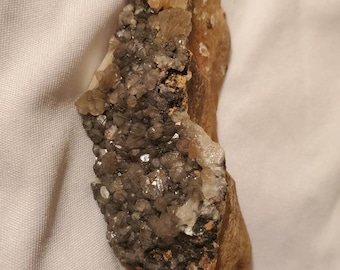 May include: A natural mineral specimen featuring a cluster of small, sparkling crystals in shades of gray and white, set against a rough, brown rock matrix. A creamy, light-colored crystal formation sits atop the rock.