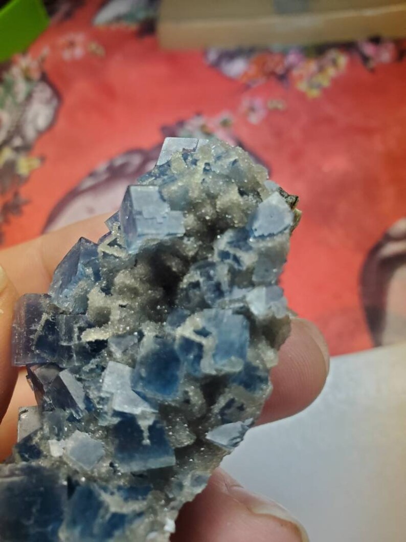 May include: Close-up of a cluster of blue and gray fluorite crystals. The crystals have a cubic shape and are arranged in a dense formation. The background is a blurred red and floral pattern.