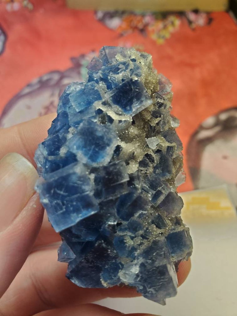 May include: A cluster of blue fluorite crystals with a cubic structure. The crystals have a translucent quality and are a deep shade of blue. Some areas have a dusting of small, light-colored crystals.