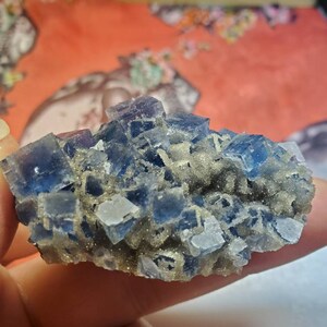 May include: A cluster of blue and clear fluorite crystals. The crystals have a cubic shape and are arranged in a layered formation. The fluorite is a deep blue color with some areas of clear transparency. The base of the cluster has a light brown, granular texture.