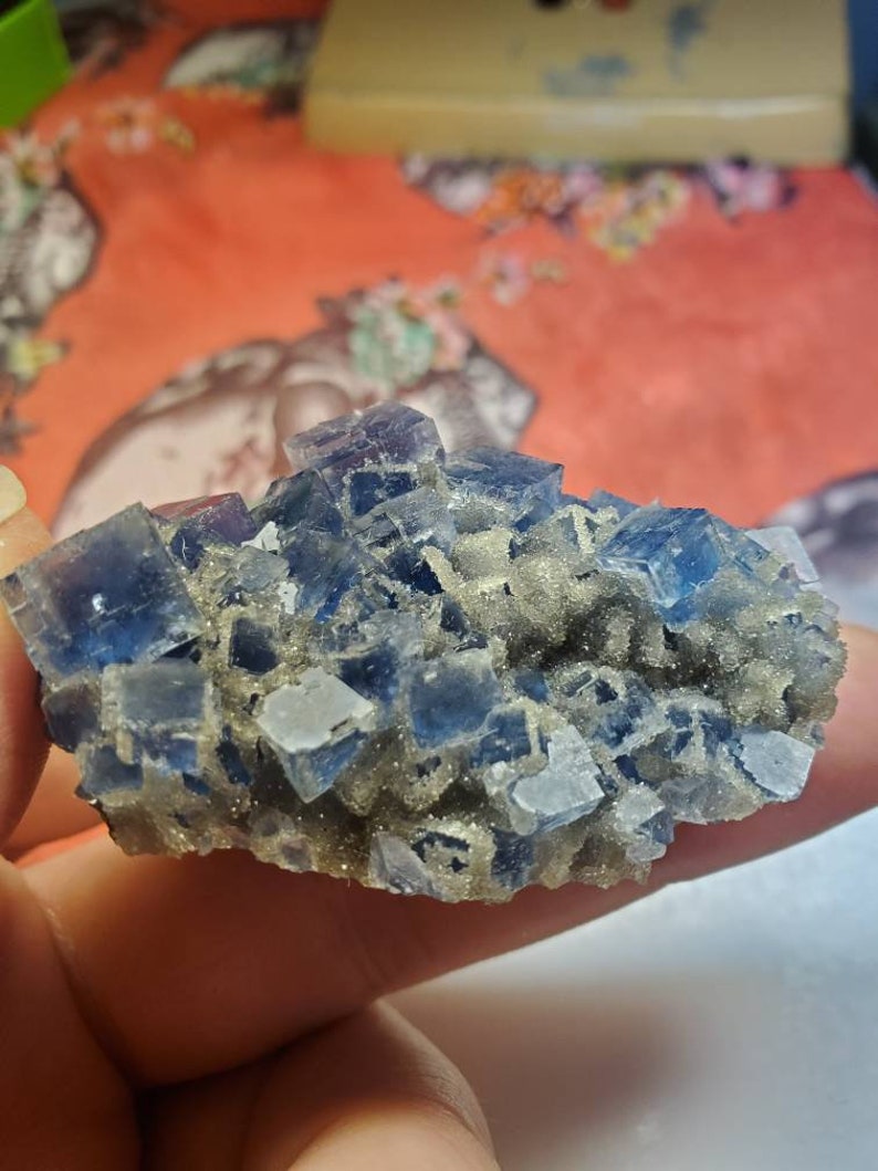 May include: A cluster of blue and clear fluorite crystals. The crystals have a cubic shape and are arranged in a layered formation. The fluorite is a deep blue color with some areas of clear transparency. The base of the cluster has a light brown, granular texture.