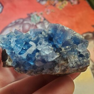 May include: A cluster of blue fluorite crystals with a rough, natural texture. The crystals are a vibrant, translucent blue, with some areas appearing more opaque. The base of the cluster is a dark, earthy brown.