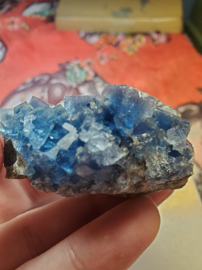 May include: A cluster of blue fluorite crystals with a rough, natural texture. The crystals are a vibrant, translucent blue, with some areas appearing more opaque. The base of the cluster is a dark, earthy brown.
