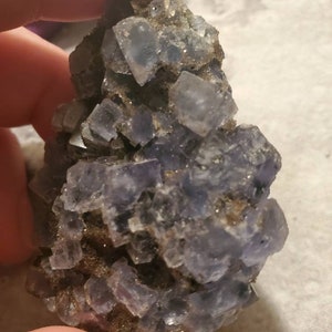 May include: A cluster of blue-purple fluorite crystals. The crystals are cubic in shape and are clustered together on a matrix of brown rock. The fluorite has a translucent appearance and reflects light. The overall appearance is rough and natural.