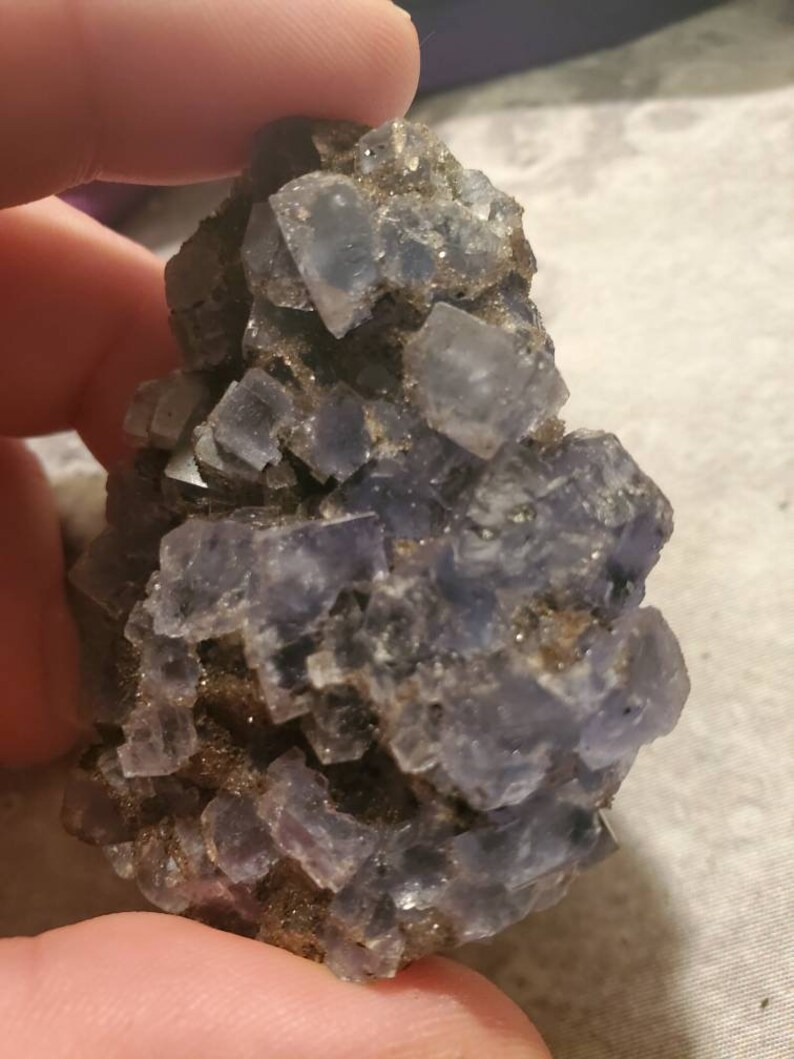 May include: A cluster of blue-purple fluorite crystals. The crystals are cubic in shape and are clustered together on a matrix of brown rock. The fluorite has a translucent appearance and reflects light. The overall appearance is rough and natural.