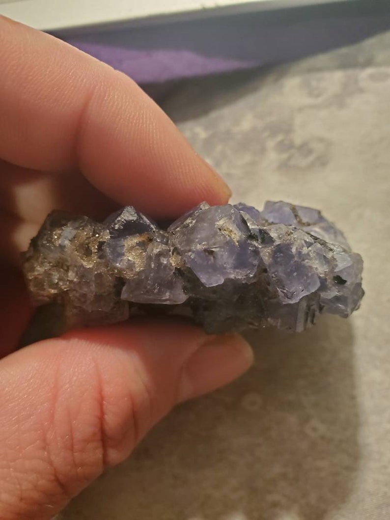 May include: A cluster of small, light purple crystals with a rough, textured surface. The crystals have a natural, irregular shape and are held in place by a dark, earthy matrix. The overall appearance is of a raw, unpolished mineral specimen.