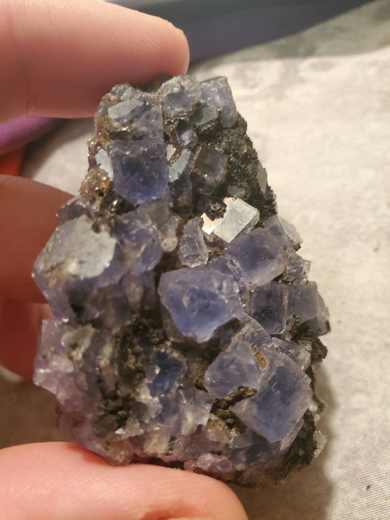 May include: A close-up of a cluster of purple fluorite crystals. The crystals are cubic in shape and have a translucent quality. The fluorite is set against a dark matrix, with some smaller, lighter-colored crystals interspersed.
