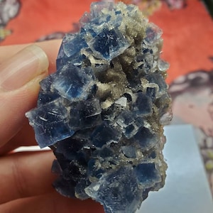 May include: A cluster of blue fluorite crystals with a cubic structure. The crystals have a deep blue color and are partially covered with a light brown, granular material. The specimen is held in a person's hand.