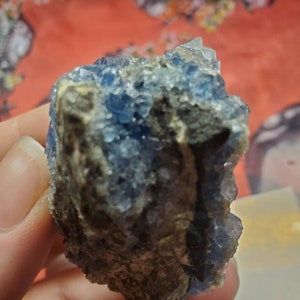 May include: A rough-hewn, natural mineral specimen featuring a cluster of small, sparkling blue crystals. The crystals are set against a backdrop of a dark, earthy-toned matrix. The overall appearance is raw and organic.