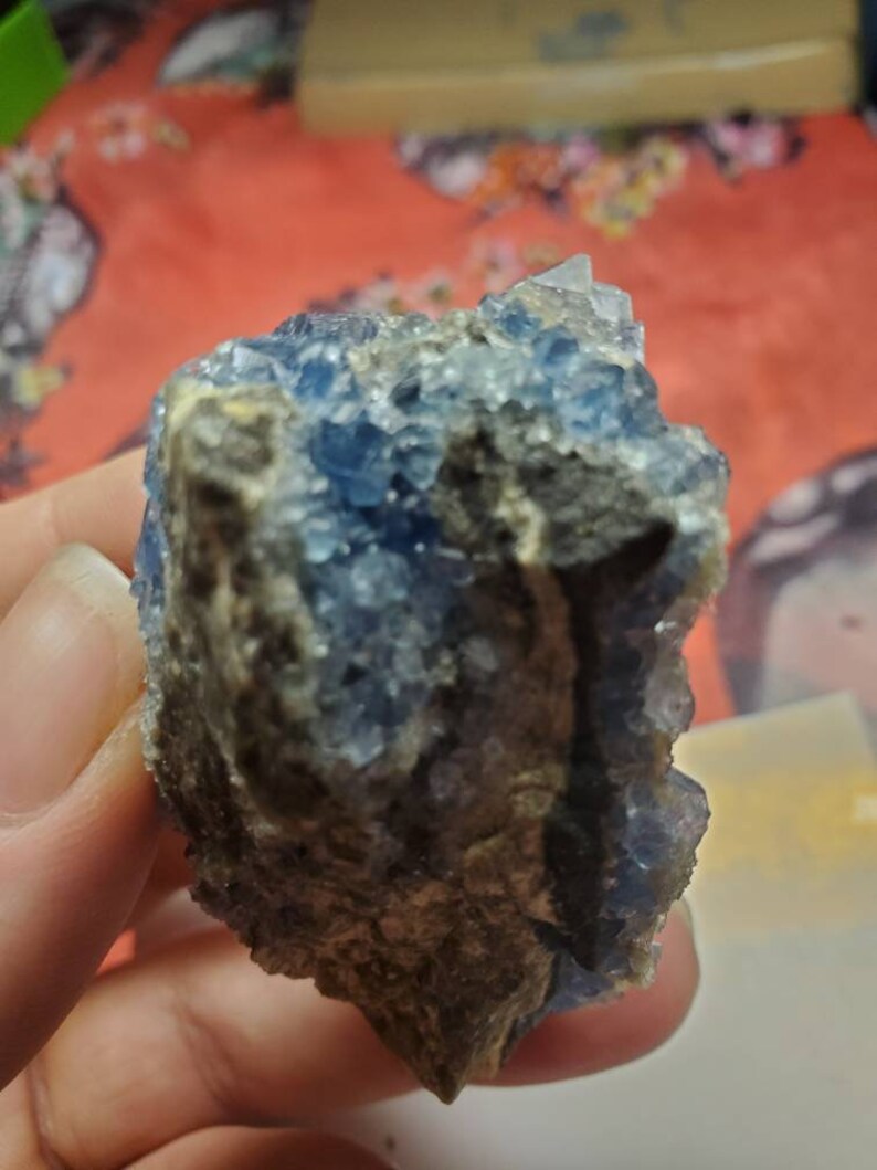May include: A rough-hewn, natural mineral specimen featuring a cluster of small, sparkling blue crystals. The crystals are set against a backdrop of a dark, earthy-toned matrix. The overall appearance is raw and organic.