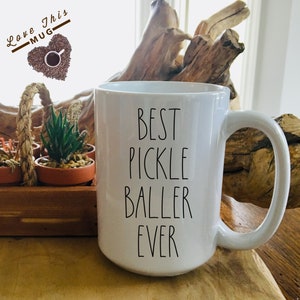 May include: White ceramic coffee mug with black text that reads "Best Pickle Baller Ever".