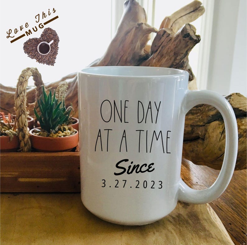 May include: White ceramic coffee mug with black text that reads "One Day At A Time Since 3.27.2023".