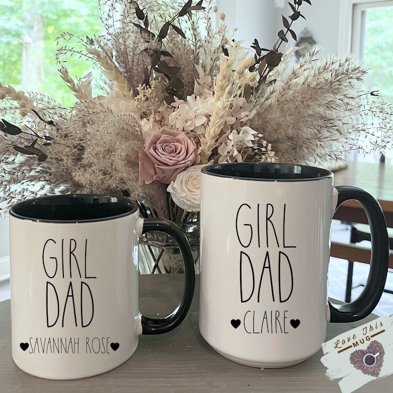 May include: Two white ceramic mugs with black handles and black rims. Both mugs have the text "Girl Dad" in black lettering. The mug on the left has the text "Savannah Rose" in black lettering with a heart on each side. The mug on the right has the text "Claire" in black lettering with a heart on each side.