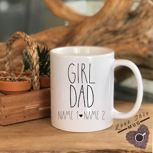 May include: White ceramic coffee mug with black text that reads "Girl Dad" and "Name 1 &hearts; Name 2". The mug has a black handle and is sitting on a wooden surface.