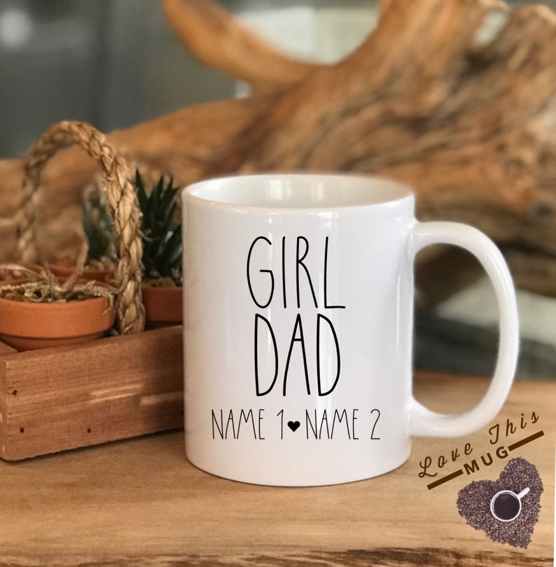 May include: White ceramic coffee mug with black text that reads "Girl Dad" and "Name 1 &hearts; Name 2". The mug has a black handle and is sitting on a wooden surface.