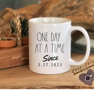 May include: White ceramic coffee mug with black text that reads "One Day At A Time Since 3.27.2023". The mug has a black heart-shaped design with the text "Love This Mug" and coffee beans.