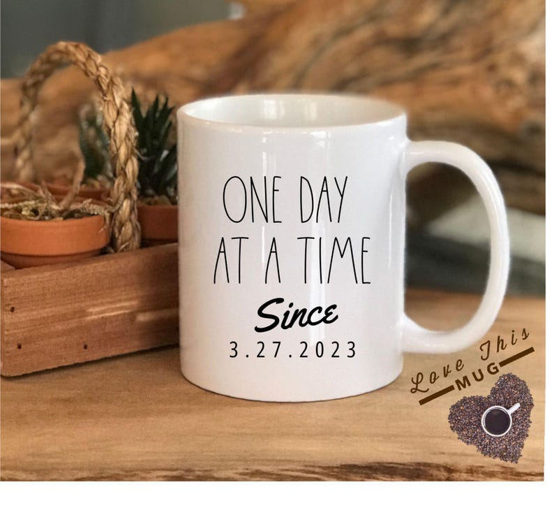 May include: White ceramic coffee mug with black text that reads "One Day At A Time Since 3.27.2023". The mug has a black heart-shaped design with the text "Love This Mug" and coffee beans.