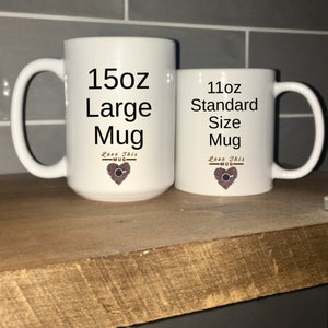 May include: Two white ceramic mugs with black text and a coffee bean heart design. The larger mug is labeled "15oz Large Mug" and the smaller mug is labeled "11oz Standard Size Mug".