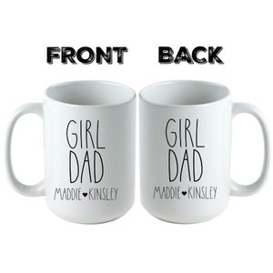 May include: Two white ceramic mugs with black text that reads "Girl Dad" and the names "Maddie" and "Kinsley" on the front and back.